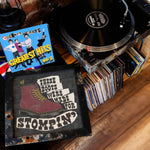 Load image into Gallery viewer, a collection of vinyl records and a framed poster with a shoe design, all placed on a wooden surface.
