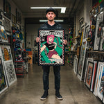 Load image into Gallery viewer, A man is standing in a room filled with various artworks, including paintings and sculptures, holding a framed poster or artwork featuring a woman with pink hair and a checkered pattern.
