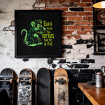 Load image into Gallery viewer, a black framed poster with a cartoon rat and text, hanging on a brick wall. There are three skateboards leaning against the wall, and a table with a can on it is visible in the foreground.
