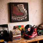 Load image into Gallery viewer, a wall with a framed poster of a red boot, a stack of CDs, a pair of red boots, and a record album.
