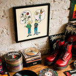 Lade das Bild in den Galerie-Viewer, a collection of various items, including a framed cartoon drawing, a stack of CDs, a pair of red boots, and a pile of vinyl records, all displayed on a wooden surface against a white brick wall.
