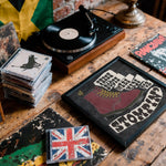 Load image into Gallery viewer, a collection of various items, including a record player, a framed poster, a stack of CDs, and a flag, all arranged on a wooden surface.
