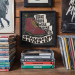 Load image into Gallery viewer, a framed poster with a graphic of a red boot, surrounded by various CDs and DVDs on a shelf.
