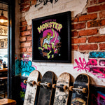 Load image into Gallery viewer, a wall with a framed poster of a cartoon monster, a row of skateboards leaning against the wall, and graffiti on the brick wall.
