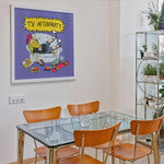 Cargar imagen en el visor de la galería, a dining room with a glass table and four wooden chairs. Above the table, there is a framed cartoon-style illustration of a person sitting on a couch with a skateboard, surrounded by various objects.
